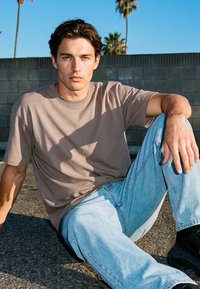 Nuori mies ruskeat hiukset, beige t-paita ja vaaleansiniset farkut, istuu jalkakäytävällä harmaata lohkokiviseinää vasten, yläpuolella palmuja.