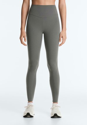 Donna che indossa leggings a vita alta color verde oliva e scarpe da ginnastica bianche, in piedi con le braccia rilassate lungo i fianchi su uno sfondo neutro.