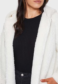 Chaqueta de forro polar blanca y esponjosa con capucha, que cuenta con una cremallera plateada y bolsillos laterales; llevada sobre una camiseta negra de canalé.