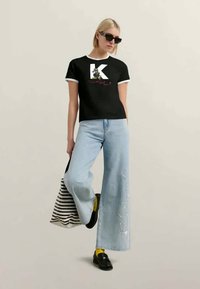 Γυναίκα με μαύρο μπλουζάκι με γραφικά, ανοιχτό μπλε φαρδιά τζιν παντελόνια με κεντήματα, κίτρινες κάλτσες, μαύρες λοφάτες, γυαλιά ηλίου, κρατώντας ριγέ τσάντα.