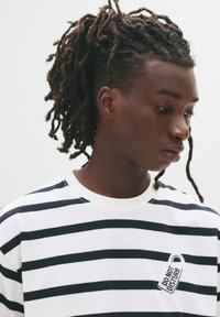 Jongere met een donkere huid en dreads, gekleed in een wit en zwart gestreept shirt met een "Niet Storen" patch, kijkend naar beneden naar rechts.