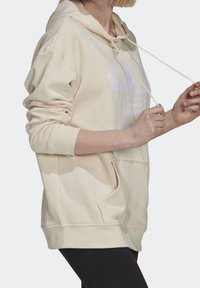 Krémově zbarvená mikina s kapucí, přední kapsou a bílým logem, vybavená stahovací kapucí a žebrovanými manžetami. Materiál působí měkký a hladký.