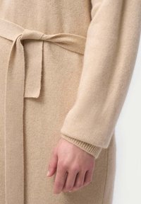 Beige kardigan med ett band i midjan, tillverkad av mjukt stickat material. Har långa ärmar, ribbade muddar och en slät yta.