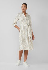 Cremfarbendes Hemdkleid mit hellbraunen Blättern, Gürtel in der Taille, Knopfleiste und dreiviertellangen Ärmeln, kombiniert mit weißen Sneakers.