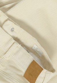 Gros plan sur la taille d'un pantalon en velours côtelé beige avec bouton, sangle élastique ajustable avec bouton, fermeture à pression et étiquette de marque rectangulaire marron.