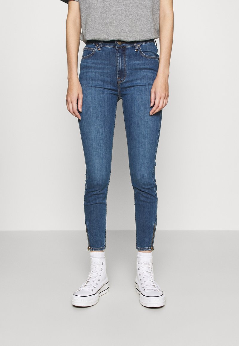 Blå denim skinny jeans med mellanhög midja, med en dragkedjedetalj vid ankeln, i kombination med vita högskurna sneakers och en grå T-shirt.
