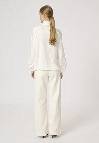 Weiße Seidenbluse mit hohem Kragen und puffigen Ärmeln, kombiniert mit weißen Hose im weiten Schnitt, was einen glatten, minimalistischen Look ergibt.