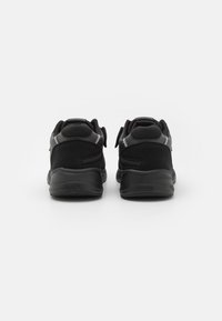 Des chaussures de sport noires présentent une combinaison de matériaux lisses et texturés, des talons arrondis et un rembourrage prononcé. Les chaussures sont dotées de sangles Velcro.