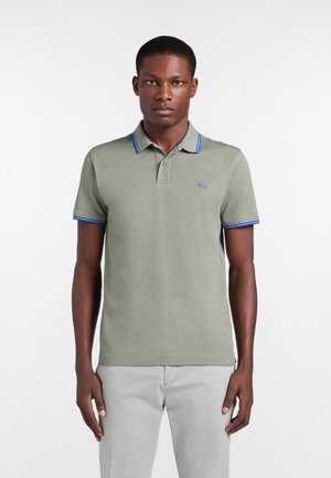 Uomo che indossa una polo grigio chiaro con colletto e maniche bordati di blu, in piedi davanti a uno sfondo bianco semplice.