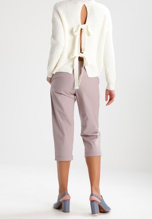 Vue de dos d’un mannequin portant un pull blanc côtelé avec des découpes dans le dos et des nœuds, un pantalon court mauve et des chaussures bleues à talon bloc.