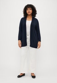 Marineblå cardigan med lange ermer og åpen front, kombinert med en hvit topp og brede hvite bukser, med en jevn tekstur.