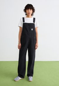 Schwarze Denim-Latzhosen mit verstellbaren Schulterträgern, frontaler Tasche und seitlichen Knöpfen. Getragen über einem weißen T-Shirt, kombiniert mit grauen Schuhen.