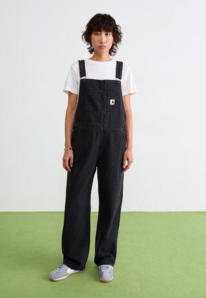 Zwarte denim overalls met verstelbare schouderbanden, een voorzak en zijknopen. Gedragen over een wit T-shirt, gecombineerd met grijze schoenen.