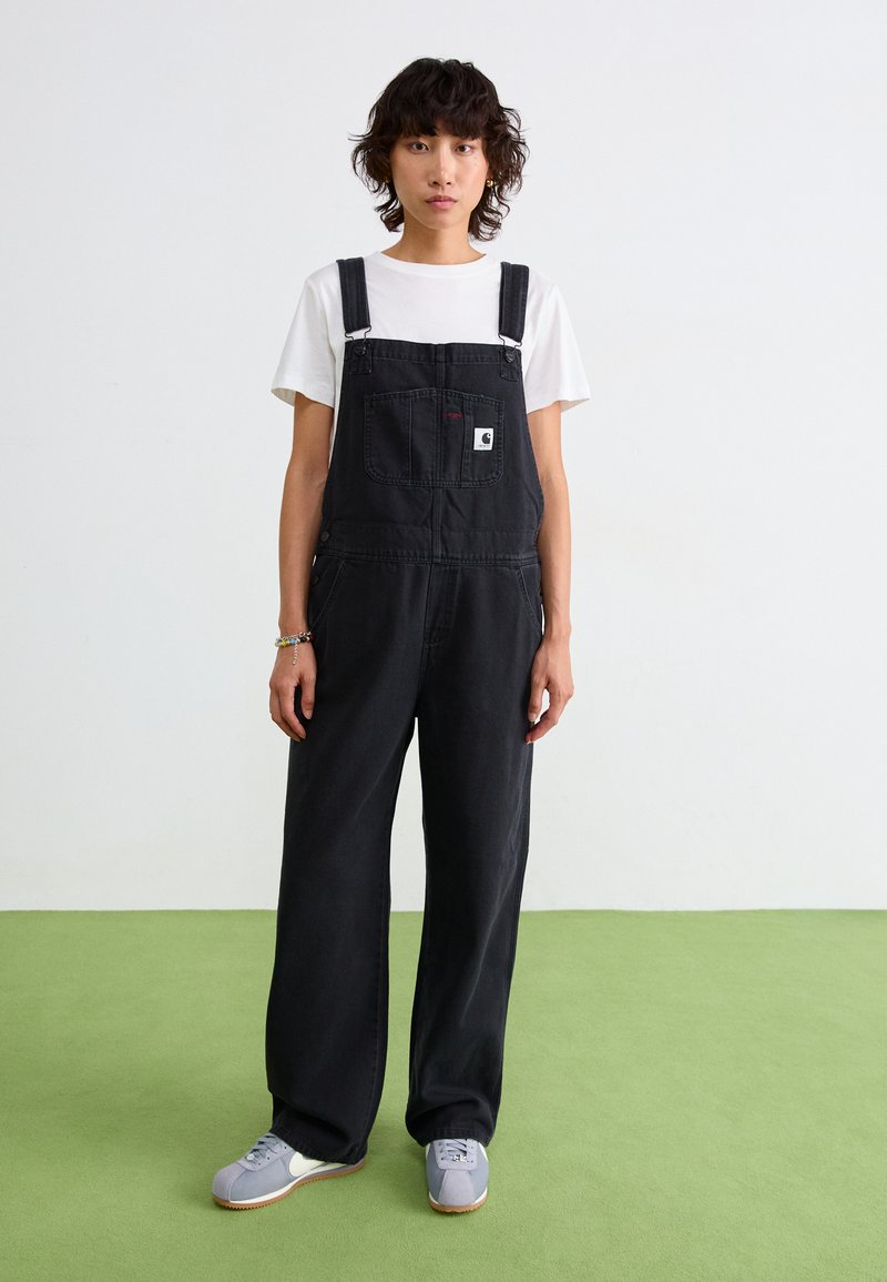 Schwarze Denim-Latzhosen mit verstellbaren Schulterträgern, frontaler Tasche und seitlichen Knöpfen. Getragen über einem weißen T-Shirt, kombiniert mit grauen Schuhen.