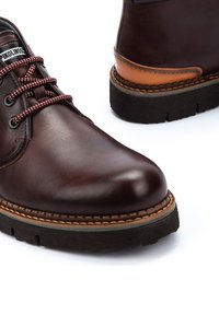 Zapatos de cuero marrón con punta redonda, detalles en beige contrastados, cordones con un patrón rojo y blanco, y una suela de goma negra texturizada.