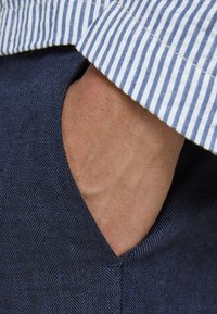 Navigátorská tkanina s tkaným vzorem herringbone, která má výřez ve tvaru písmene V pro kapsu a světlomodrý pruhovaný límec.