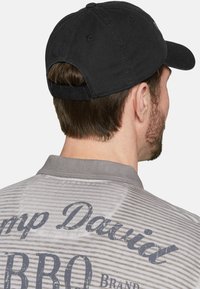 Schwarze Baseballmütze mit gebogener Krempe, aus Baumwolle, rückwärts getragen, zusammen mit einem grauen gestreiften Hemd, auf dessen Rücken blauer Text steht.