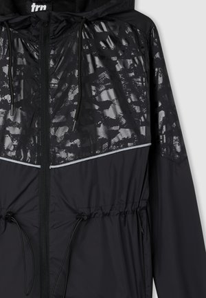 Giacca a vento nera con parte superiore lucida a motivo nero, vita regolabile con cordino e dettagli riflettenti lungo la cucitura del petto.