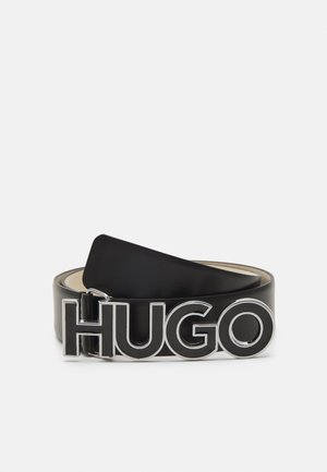 Schwarzer Ledergürtel zusammengerollt mit großen silberumrandeten Buchstaben, die "HUGO" als Schnalle auf weißem Hintergrund formen.