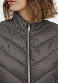 Pilkas pūkinis švarkas su aukštu apykakle ir chevrono modelio siuvinėjimu. Yra priekinis užtrauktukas ir blizgus, lygus paviršius.