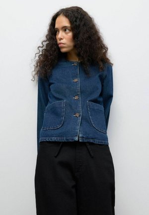 Denimjakke med knaplukning, to store lommer, lange ærmer og en blå nuance, parret med sorte bukser. Glat tekstur og struktureret design.