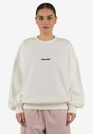 Vrouw met donker haar in een staart draagt een losse witte trui met de tekst "PEGADOR" en lichtroze broek tegen een eenvoudige lichte achtergrond.