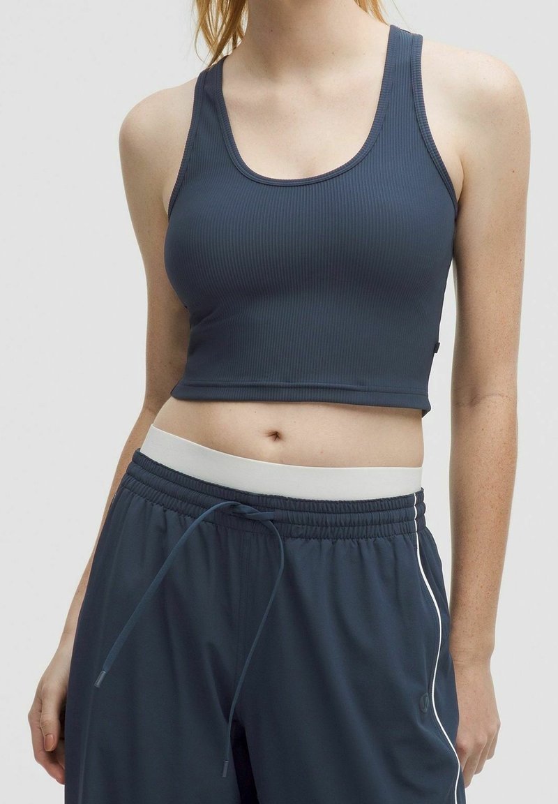 Femme portant un débardeur court côtelé bleu foncé et un pantalon de sport bleu marine à cordon avec une ceinture blanche et une bande latérale.