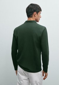 Donkergroen poloshirt met lange mouwen, een klassieke kraag, naadloze schouders en een zachte textuur, gedragen met witte broek.