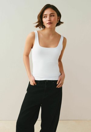 Rövid barna hajú nő, fehér ujjatlan felsőben és bő fekete nadrágban, kezét zsebében tartva, oldalra néz egy sima háttér előtt.