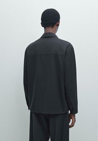 Giacca nera con colletto, maniche lunghe, caratterizzata da una texture liscia e un taglio dritto. Dettagli con bottoni sui polsini, design minimalista.