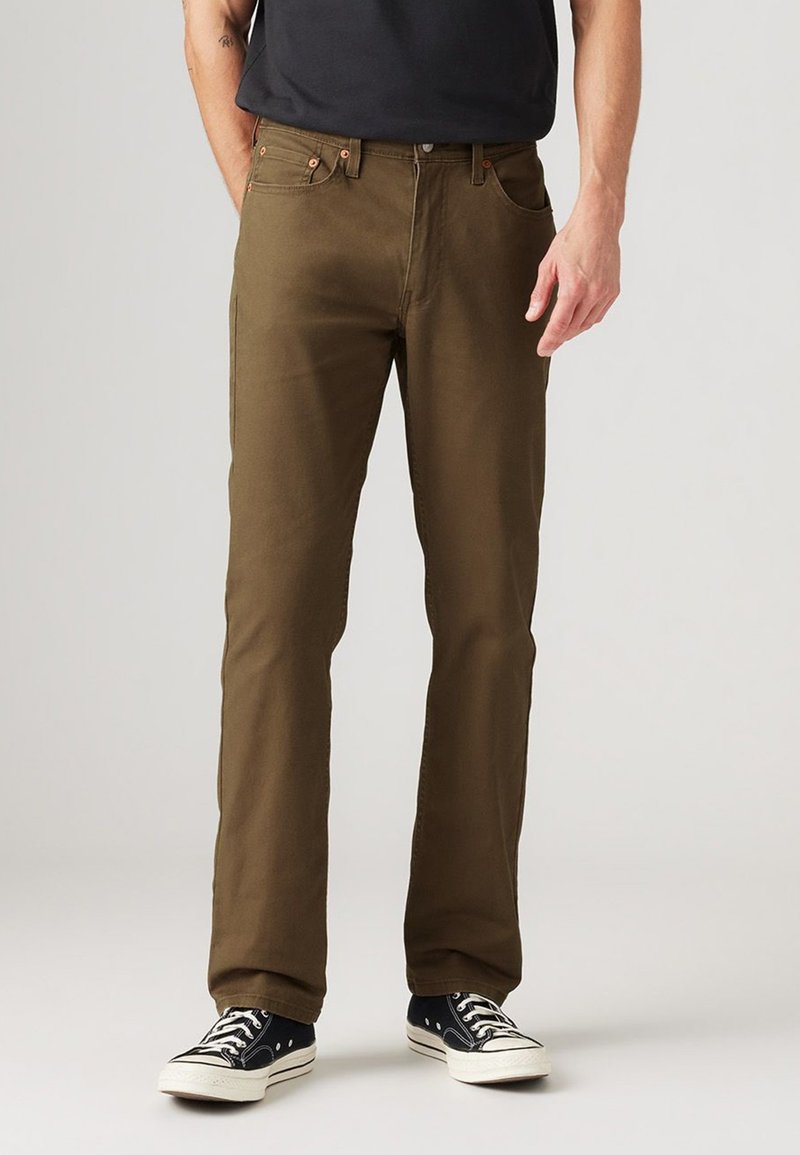 Brown cotton pants with a straight fit, featuring five pockets, contrasting stitching, and metal hardware. Paired with black sneakers.