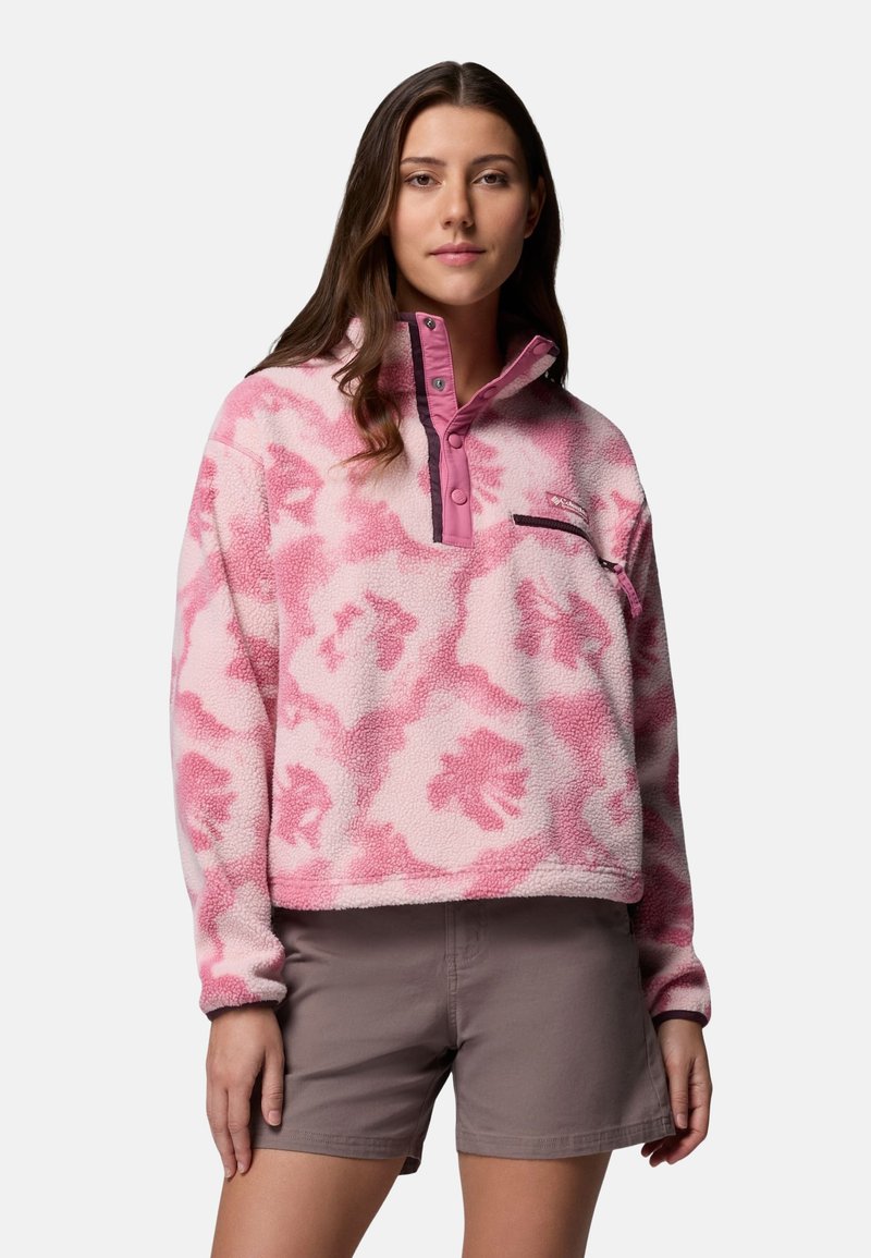 Jeune femme portant un pull polaire à motifs roses avec boutons-pression et un short taupe, posant devant un fond uni.