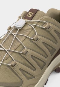 Μπεζ και καφέ παπούτσι πεζοπορίας με άνω μέρος από διχτυωτό υλικό, συνθετικές επικαλύψεις, σύστημα κορδονιών γρήγορης σύσφιξης και μαλακή σόλα για υπαίθρια χρήση.