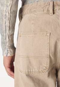 Närbild av en bakficka på beiga byxor med en hand som vilar bredvid, samt en gråbeige långärmad ribbad tröja som syns på armen.