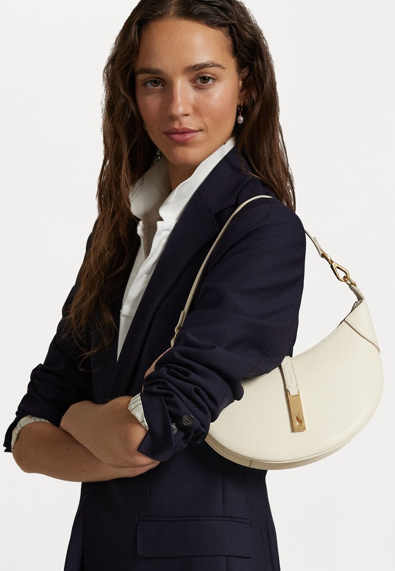 Λευκή δερμάτινη τσάντα με καμπύλο σχήμα, χρυσά αξεσουάρ και υφή, φορεμένη πάνω από ένα σκούρο σακάκι και λευκό πουκάμισο.