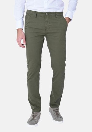 Uomo che indossa pantaloni aderenti verde oliva, camicia bianca infilata e sneakers grigie, in piedi con la mano sinistra in tasca.