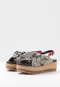 Sandales compensées avec des nœuds en tissu à motifs noir et blanc, des brides de cheville rouges réglables et des semelles compensées tissées avec un dessous marron texturé.