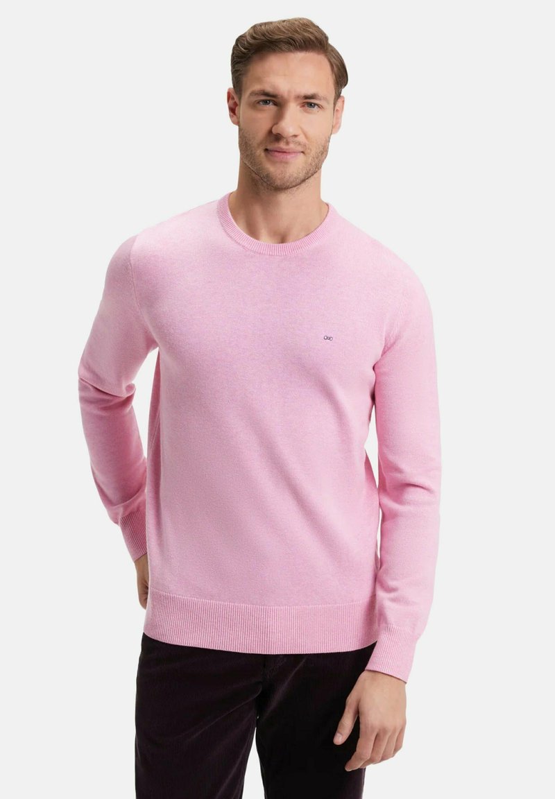 Homme portant un pull ras du cou rose clair et un pantalon sombre, se tenant avec une main dans le dos sur fond blanc.