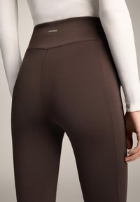 Hoge taille, bruine legging gemaakt van rekbaar materiaal. Bevat een zachte textuur en een klein logo bij de tailleband. Stikseldetails zijn zichtbaar.