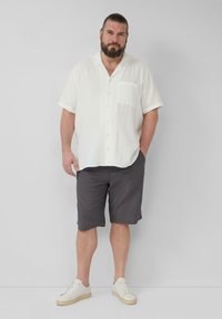 Korte mouwen witte linnen shirt met knopen en zak, gecombineerd met donkergekleurde shorts met patroon en witte sneakers, staande voor een neutrale achtergrond.