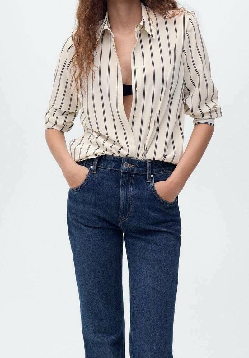 Personne portant un jean bleu taille haute et une chemise crème à boutons avec des rayures verticales noires, mains dans les poches, chemise partiellement ouverte.