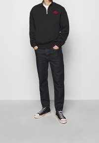 Black zip-up sweatshirt with red logo, paired with dark blue jeans and black sneakers with white soles and laces. Casual outfit presentation.