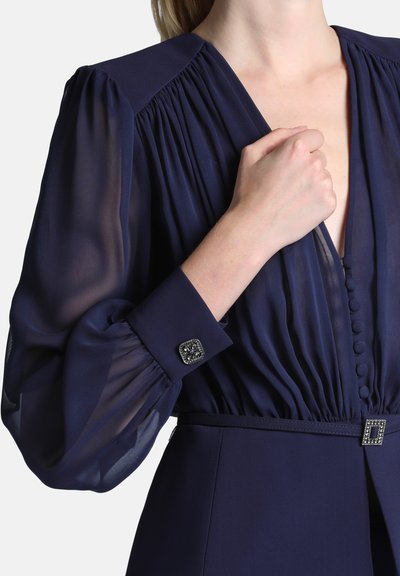 Femme portant une robe bleu marine avec de longues manches bouffantes transparentes, des boutons recouverts de tissu sur le devant et des accents décoratifs ornés de bijoux aux poignets et à la taille.