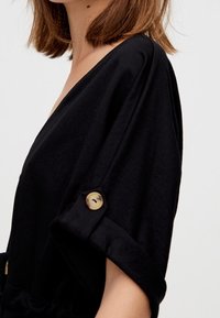 Czarny strój z głębokim dekoltem w kształcie litery V i szerokimi rękawami. Na mankietach rękawów znajdują się guziki. Wykonany z gładkiej tkaniny, dopasowany w talii.