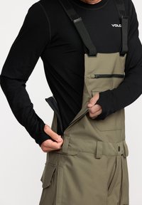 Kaki bretels met verstelbare zwarte banden, ritszak op de borst en zij-rits detail, gedragen over een zwart langemouwen shirt.