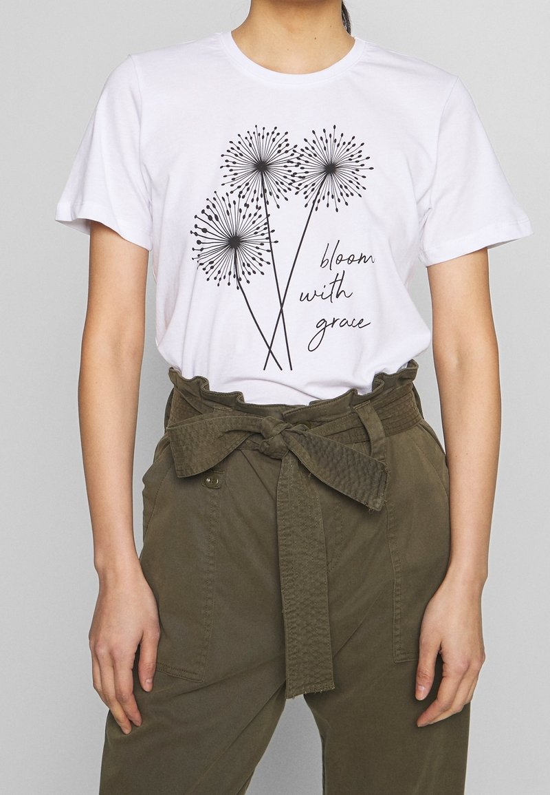 Camiseta de algodón blanca con un gráfico de diente de león negro y el texto "florece con gracia", combinada con pantalones de tiro alto verde oliva con un cinturón atado.