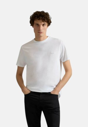 Giovane uomo con capelli ricci che indossa una semplice maglietta bianca e pantaloni neri, in piedi con le mani in tasca davanti a uno sfondo bianco.