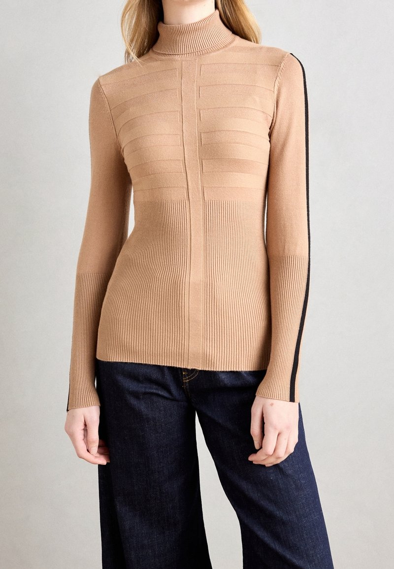 Pull à col roulé côtelé beige avec des rayures horizontales texturées et une bande latérale noire, accompagné d'un jean bleu foncé à jambes larges.