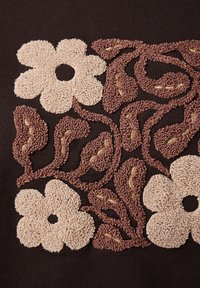 Geëmbroideerde textuur van drie beige bloemen en verschillende bruine bladeren met gele draadaccenten op een donkere stofachtergrond.