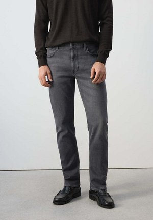 Mann trägt dunkelgraue Slim-Fit-Jeans, schwarze Loafers und einen dunklen Langarm-Pullover und steht auf einem hellgrauen Boden.